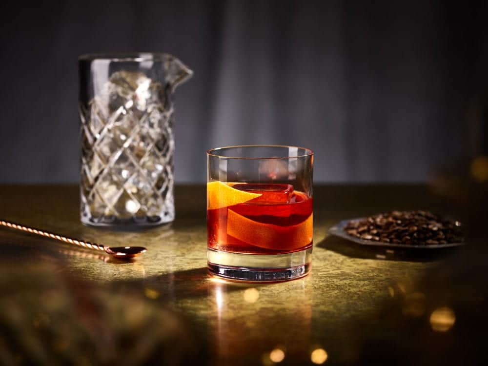 un pahar de negroni este asezat in fata unei carafe de cristai și a unei tăvi cu boabe de cafea