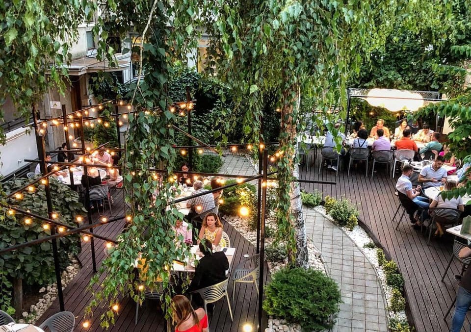 Terasa Savart din București, fotografiată de sus, cu oameni asezati la mese, copaci inalti si arbusti ornamentali, alee si beculete deasupra meselor