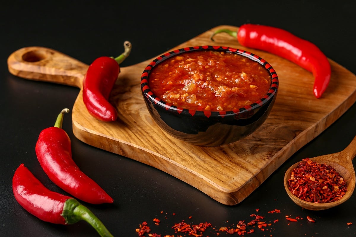 Pasta harissa, sos magrebian traditional, cu sos chili, in bol negru, fotografiat pe tocator de lemn, alaturi de ardei iuti intregi, pe fundal negru