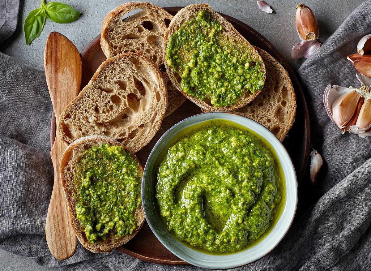 sos pesto intr-un bol alb, asezat pe un tocator rotund de lemn, alaturi de felii de paine taiate, unele unse cu sos pesto, fotografit de sus. Unul dintre cele mai populare sosuri din lume