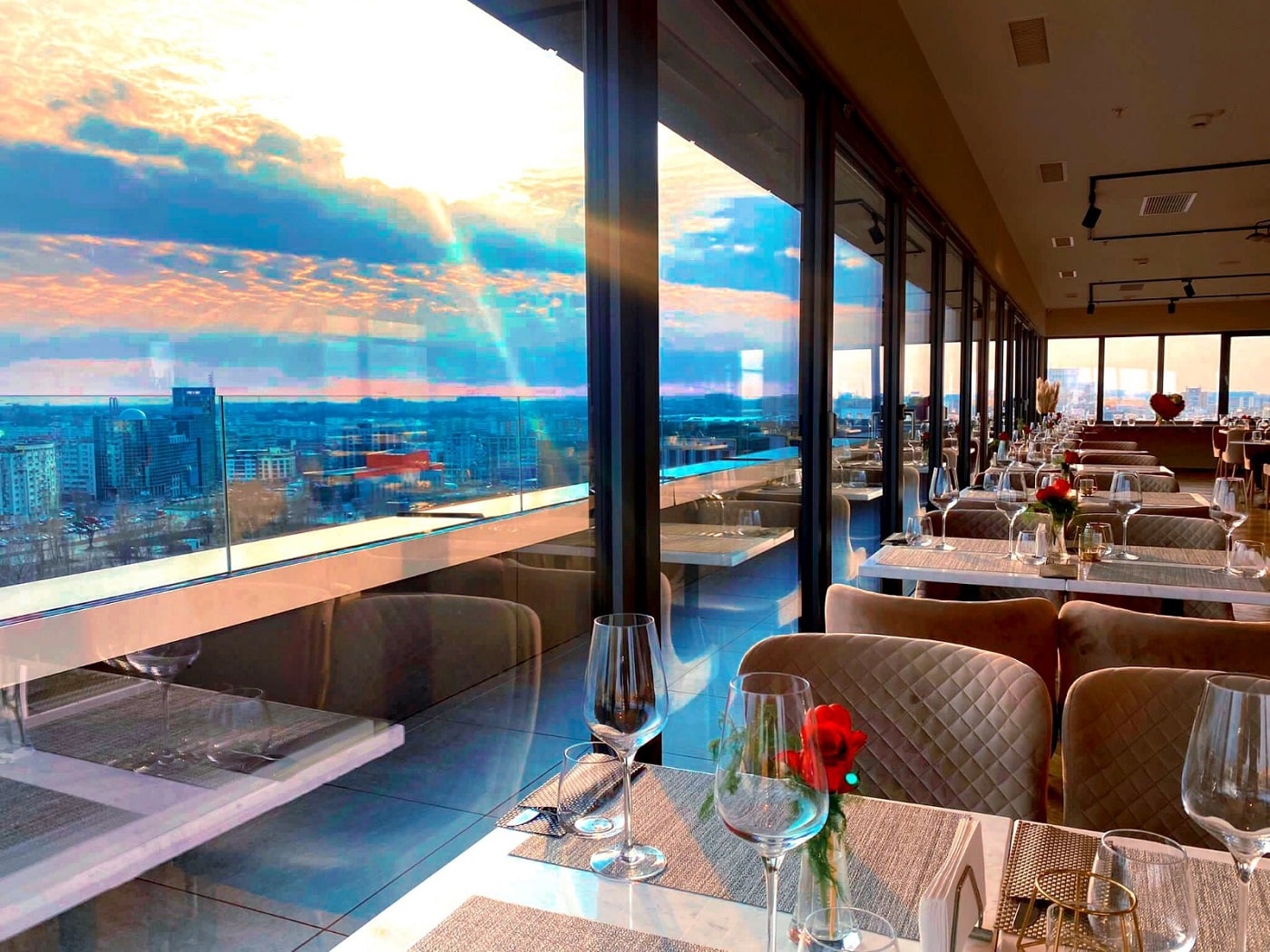 Interiorul restaurantului Amethyst Sky Bar, unul dintre restaurante romantice Bucure;ti, cu mese aranjate cu pahare si stergare si ferestre mari prin care se vede apusul