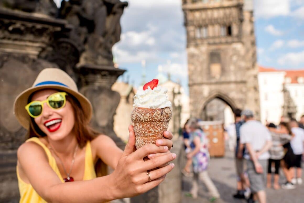 o femeie cu palarie si ochelari de soare tine in mana un TRDELNIK cu umplutura de frisca si jumatate dfe capsuna pe o strada din praga