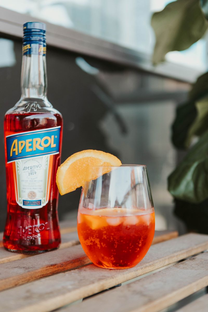un pahar de aperol spritz decorat cu o felie de portocala și o sticla de aperol pe o masa de lemn