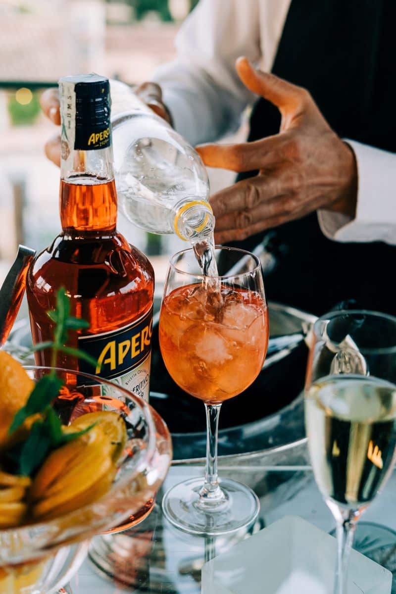 un barman pregătește un pahar de aperol spritz turnând apa minerala peste prosecco și aperol. Imagine sugestiva pentru Aperol vs Campari