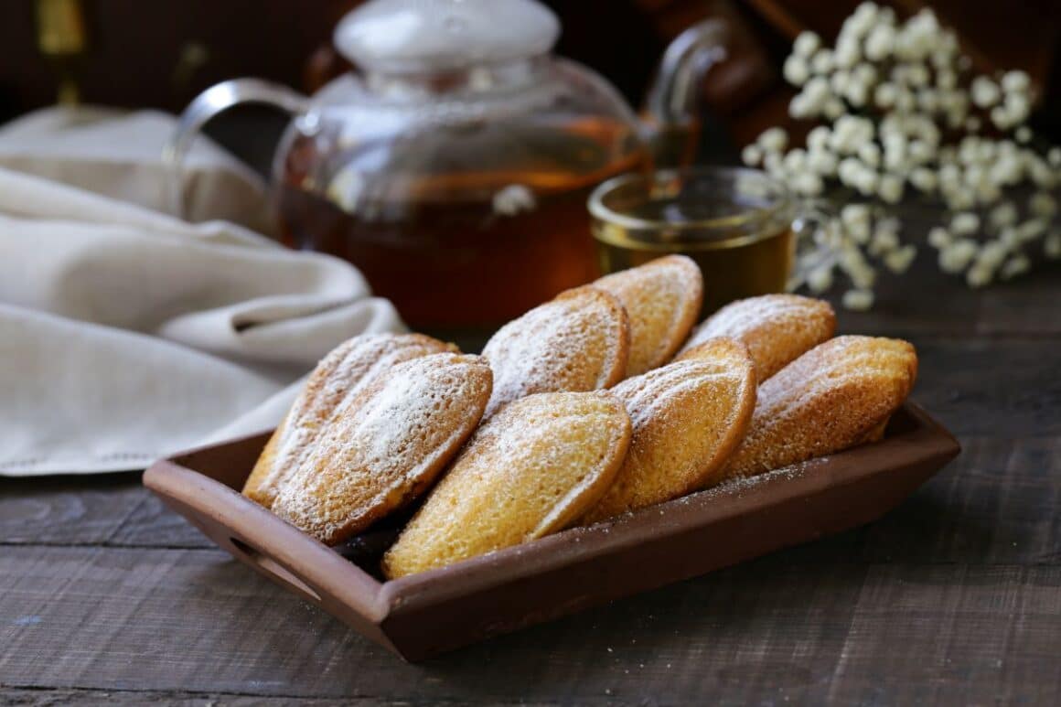 o tava de madlene este asezata in fata unui ceainic si unui pahar de ceai pe o masa de lemn