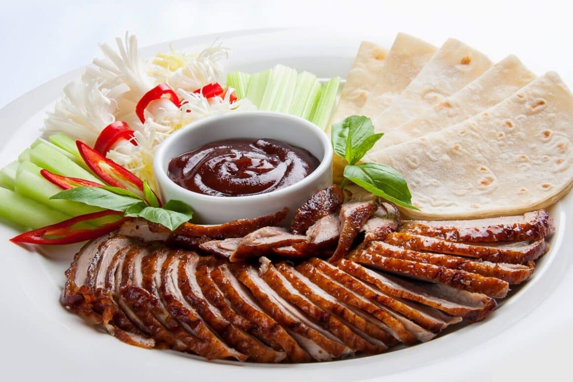 O portie de piept de rata peking este servita alaturi de castravete, praz, tije de apio și lipii pe un platou alb si cu un bol de sos in mijloc