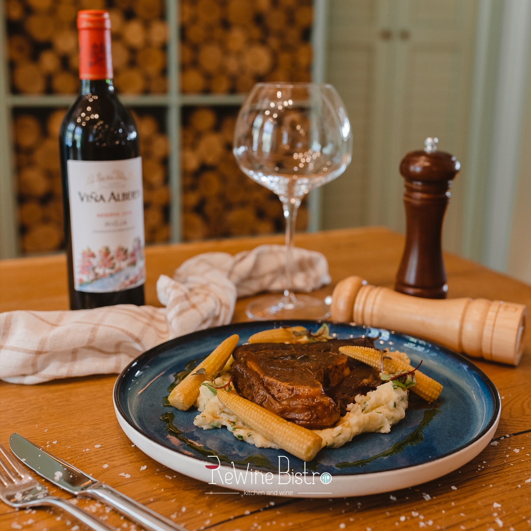 farfurie albastra cu steak de vita cu piure si morcovi, pe masa langa o sticla si un pahar de vin la Rewine bistro