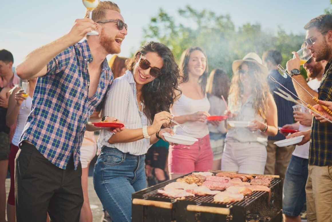mai multi tineri se distrează în jurul unui gratar fumegand, cu pahare și farfurii in mana