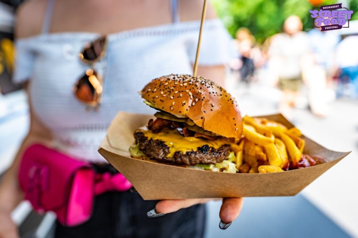 Prim plan cu un burger si cartofi prajiti intr-o cutie de carton, iar in fundal femeie care il tine in mana la festivalul de street food din Bucuresti pe Kiseleff