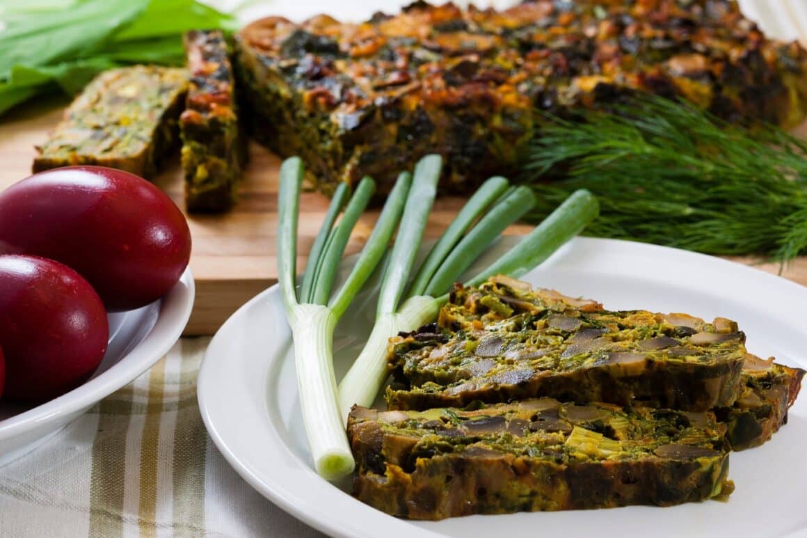 Rețete de Paști: felii de drob asezate pe o farfurie alba langa 2 fire de ceapa verde, langa o farfurie cu oua rosii. Drob gătit după rețeta tradiționlă a ui Radu Anton Roman