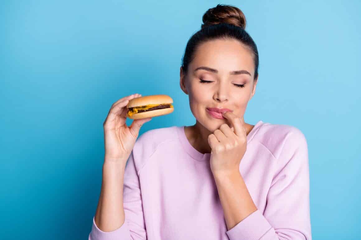 Portret close-up al unei femei care tine in mana un burger si se linge pe degete, fotografiata pe fundal albastru. Imagine concept pentru alimente care taie foamea