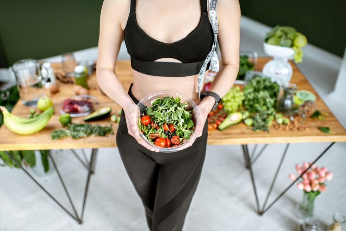 o femeie tine in mana un bol de legume, iar in spate este o masa plina de legume si verdeturi