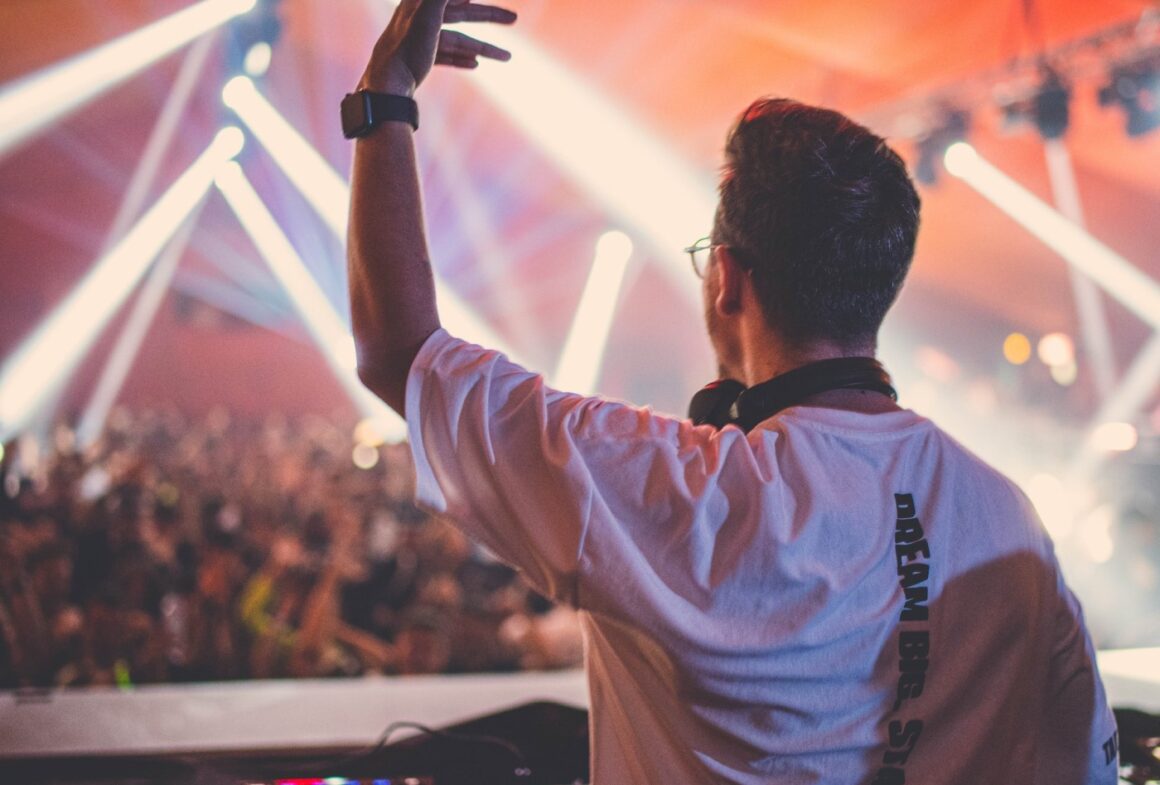 prim plan cu un dj fotografiat din spate, iar in fundal multimea de oameni si reflectoare, blurat