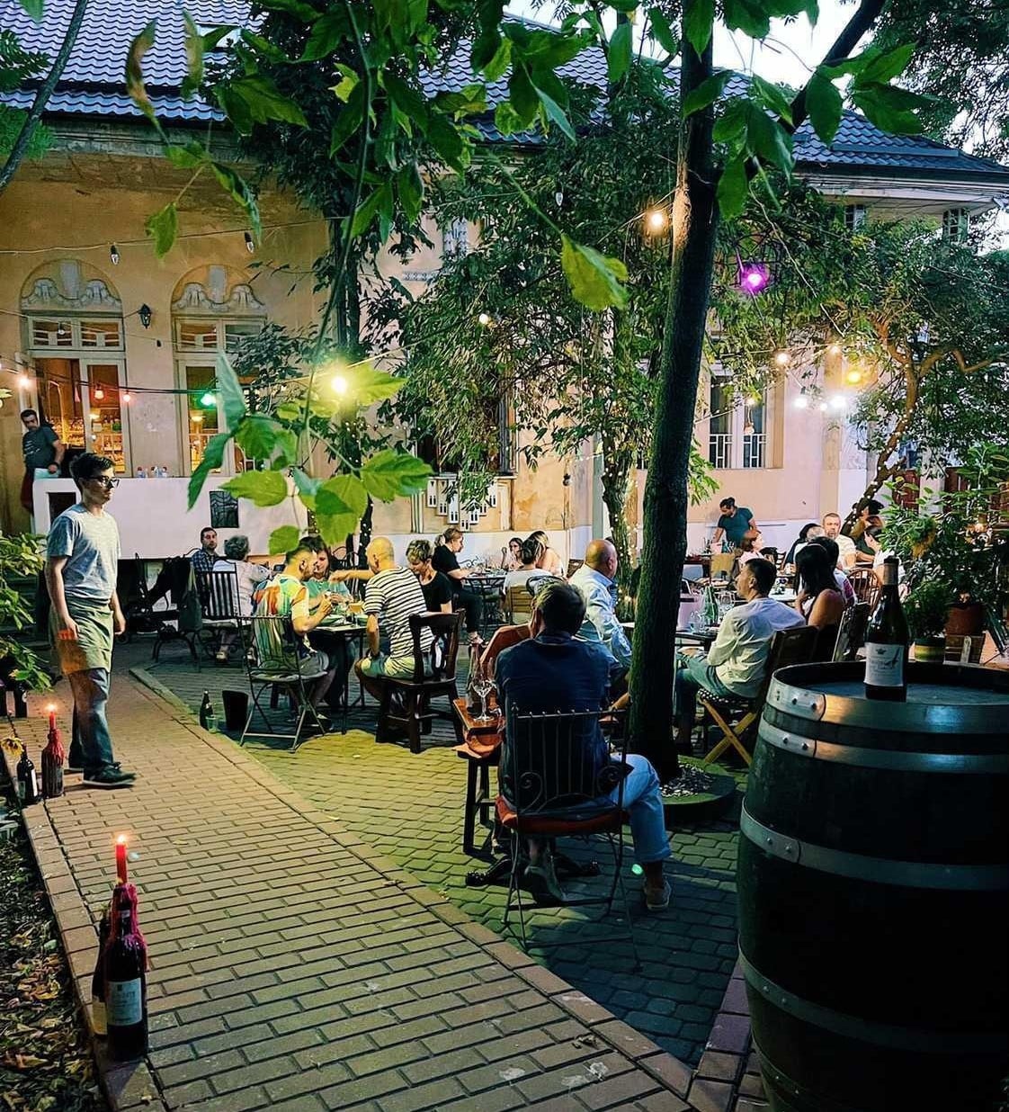 terasa Voiala Bistro, cu oameni asezati la mesele aflate printre copaci. Unde ieșim la restaurant de 1 Mai