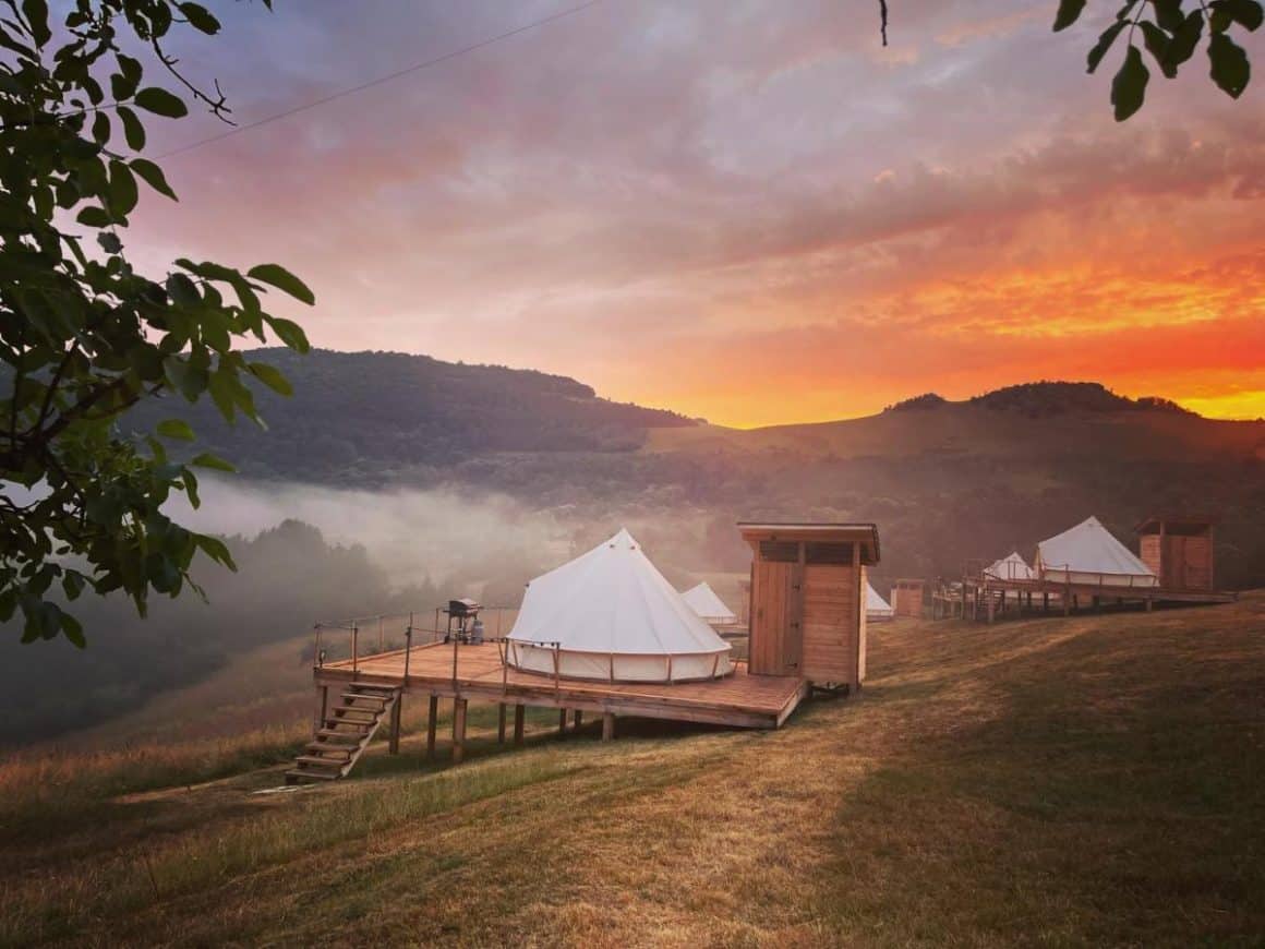 corturi de glamping rasfirate pe o pajiste, amplasate pe structura de lemn, fotografiate seara la apus la munte la The Hundetrs Glamping. Glamping în România