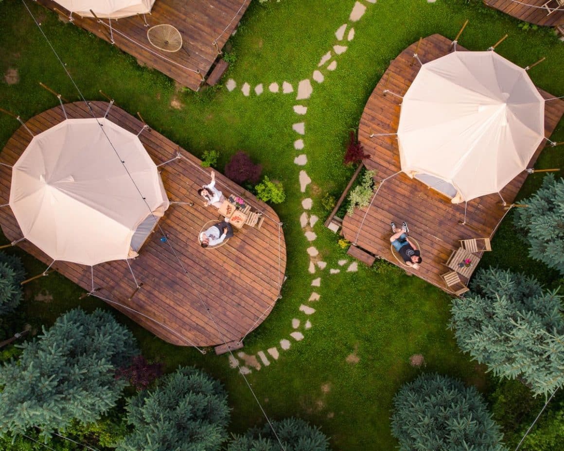 VEDERE AERIANA CU DOUA TERASE PE CARE SUNT AMPLASATE CATE UN CORT, IAR O PERSOANA STA PE VERANDA. Glamping în românia