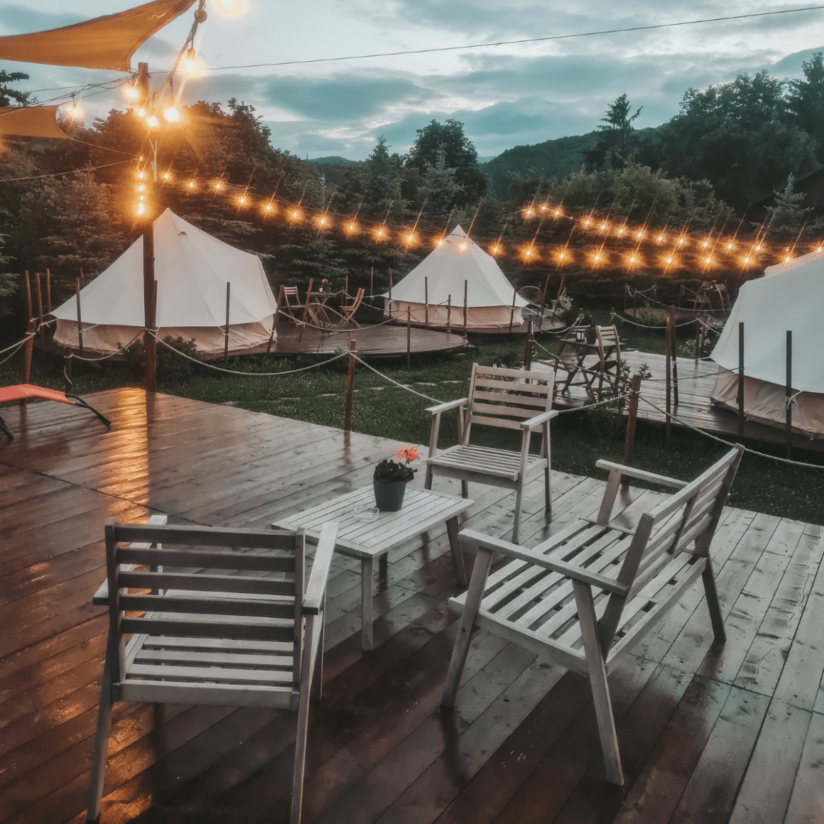 Complexul de glamping în România Valea Doftanei Glamping, cu mai multe corturi asezate pe gazon si marginite de copaci, cu luminite aprinse printre ele