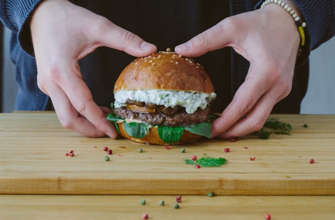 un barbat tine in maini un burger asezat pe un tocator de lemn, fotografiat de aproape. Burger Ala de miel de la Furgoneta. Burgeri gustoși în București