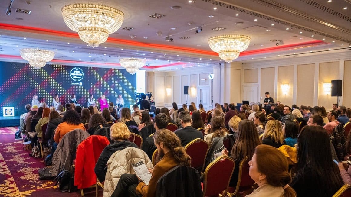 Mai multi oameni aseazati pe scaune intr-o sala de conferinte, iar in fundal o scena cu mai multi vorbitori la Food Bloggers Conference