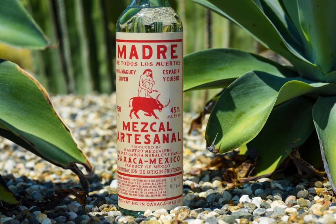 sticla de mezcal cu eticheta alba si scris rosu, fotografiata pe pamant printre frunze de agave. Ce este mezcalul