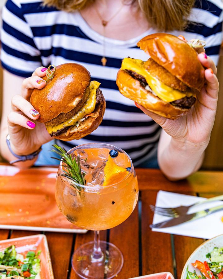 femeie tine in maini doi burgeri de la Hard Rock Cafe si in fata are un pahar de cockatil