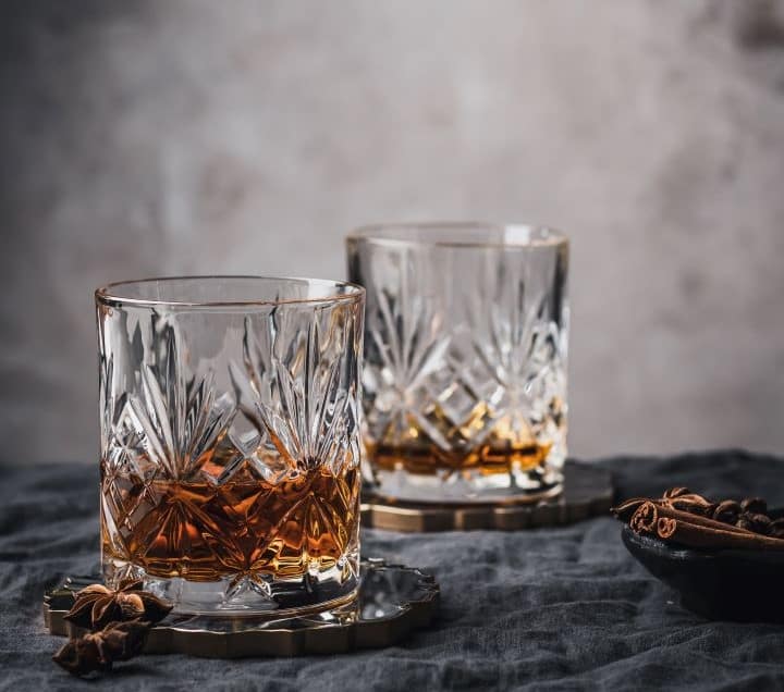 doua pahare rocks in care este putin whiskey sunt asezate pe un fundal gri, alaturi de cateva semite de anason stelat. Imagine pentru forma paharului de cocktail Rocks