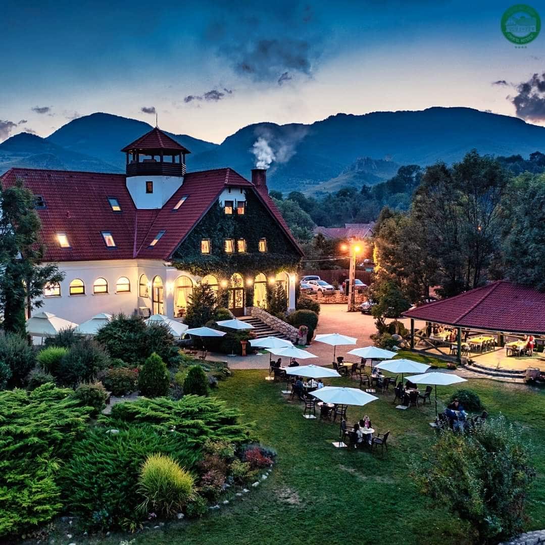 Conacul Secuiesc si curtea din fata unde sunt asezate mai multe mese, sub umbrelute, fotografiate seara. Restaurante de la sate, România culinară tradițională