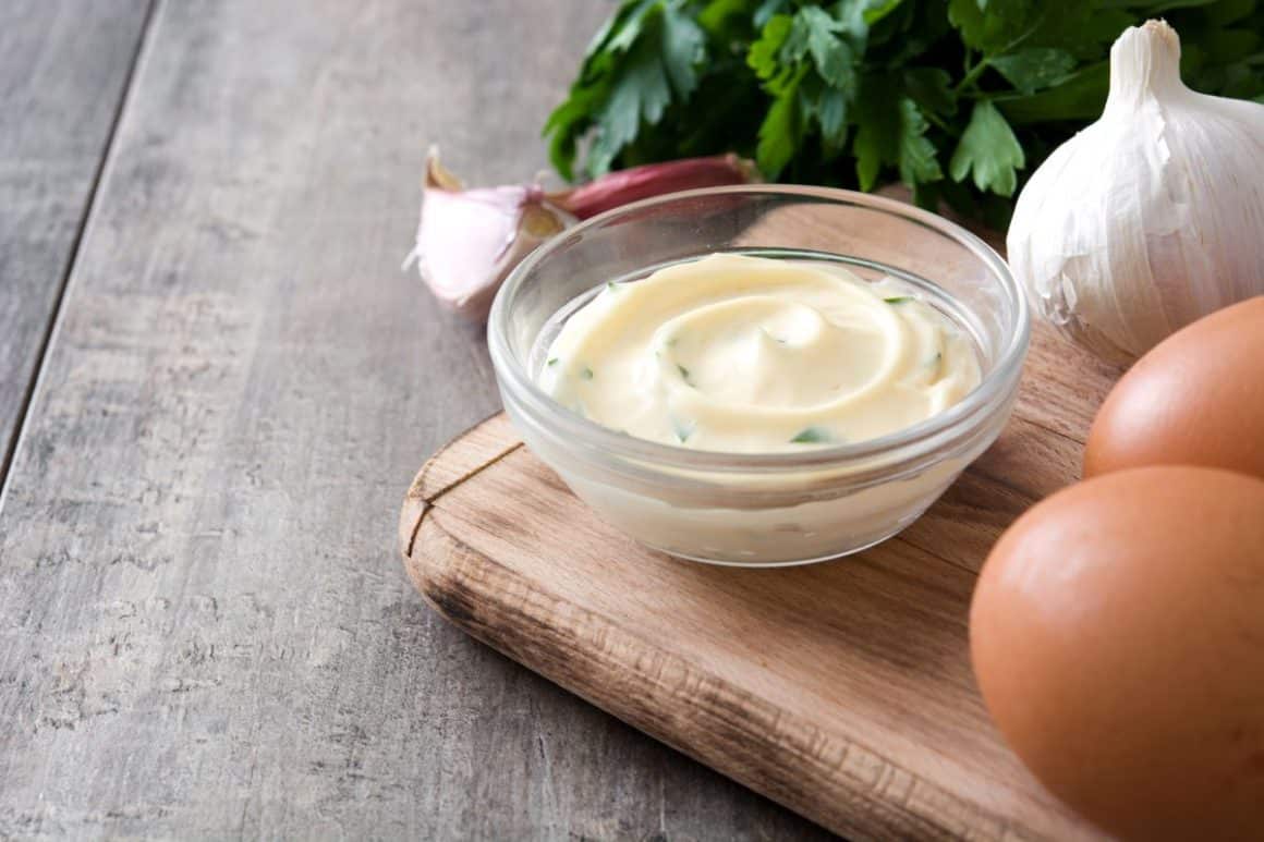 un recipient cu sosul alioli este asezat pe un fund de bucătărie, alaturi de usturoi și două ouă