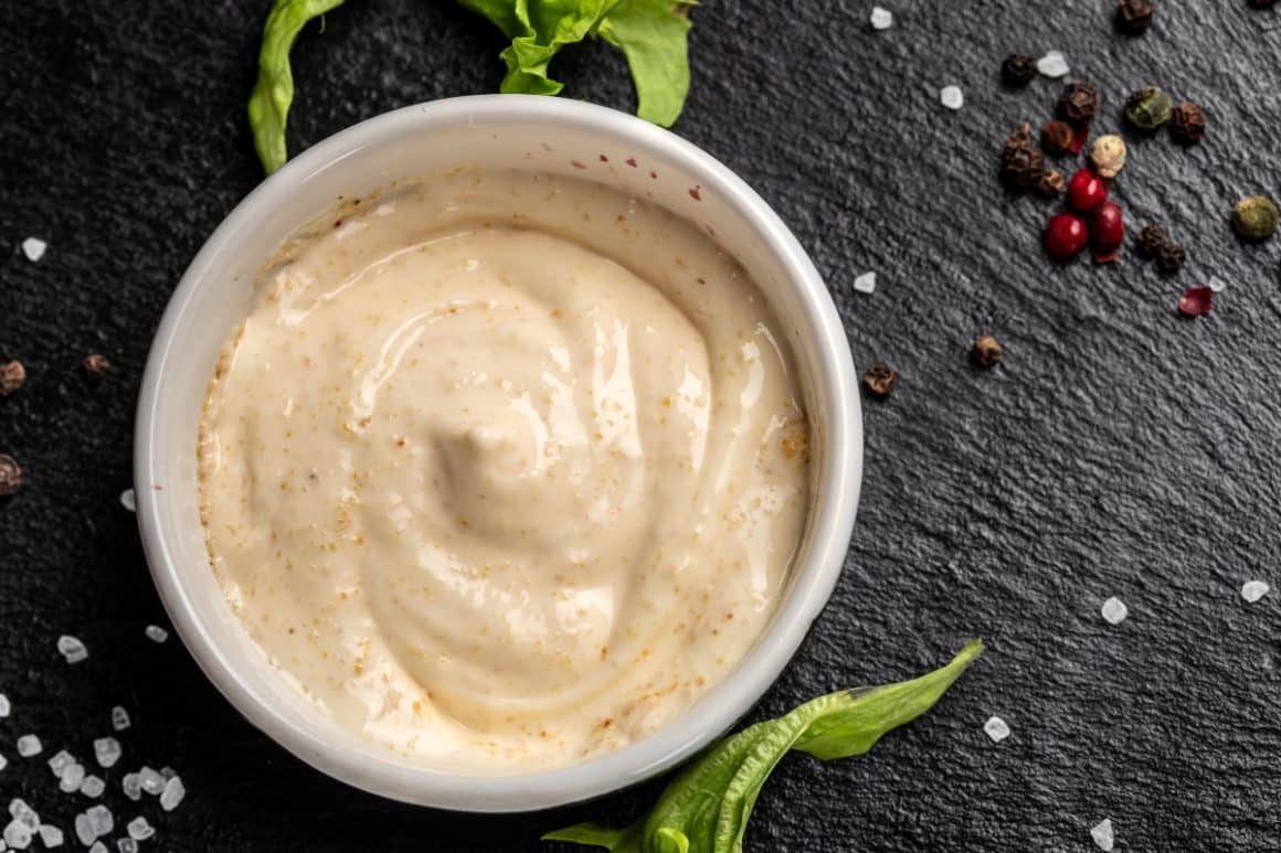 o portie de alioli intr-un bol alb, pus pe un suport negru alaturi de boabe de piper rosu și sare de mare