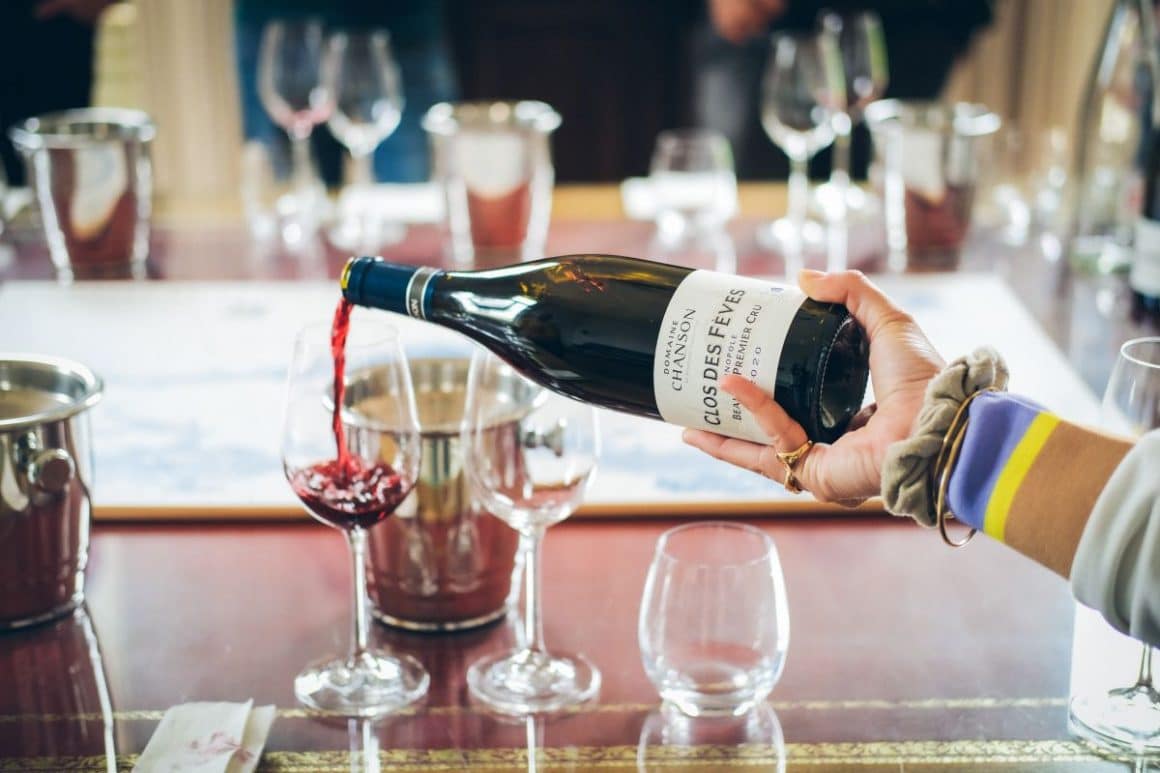 o femeie toarnă vin intr-un pahar, asezate pe o masă de lemn cu o frapieră și mai multe pahare goale