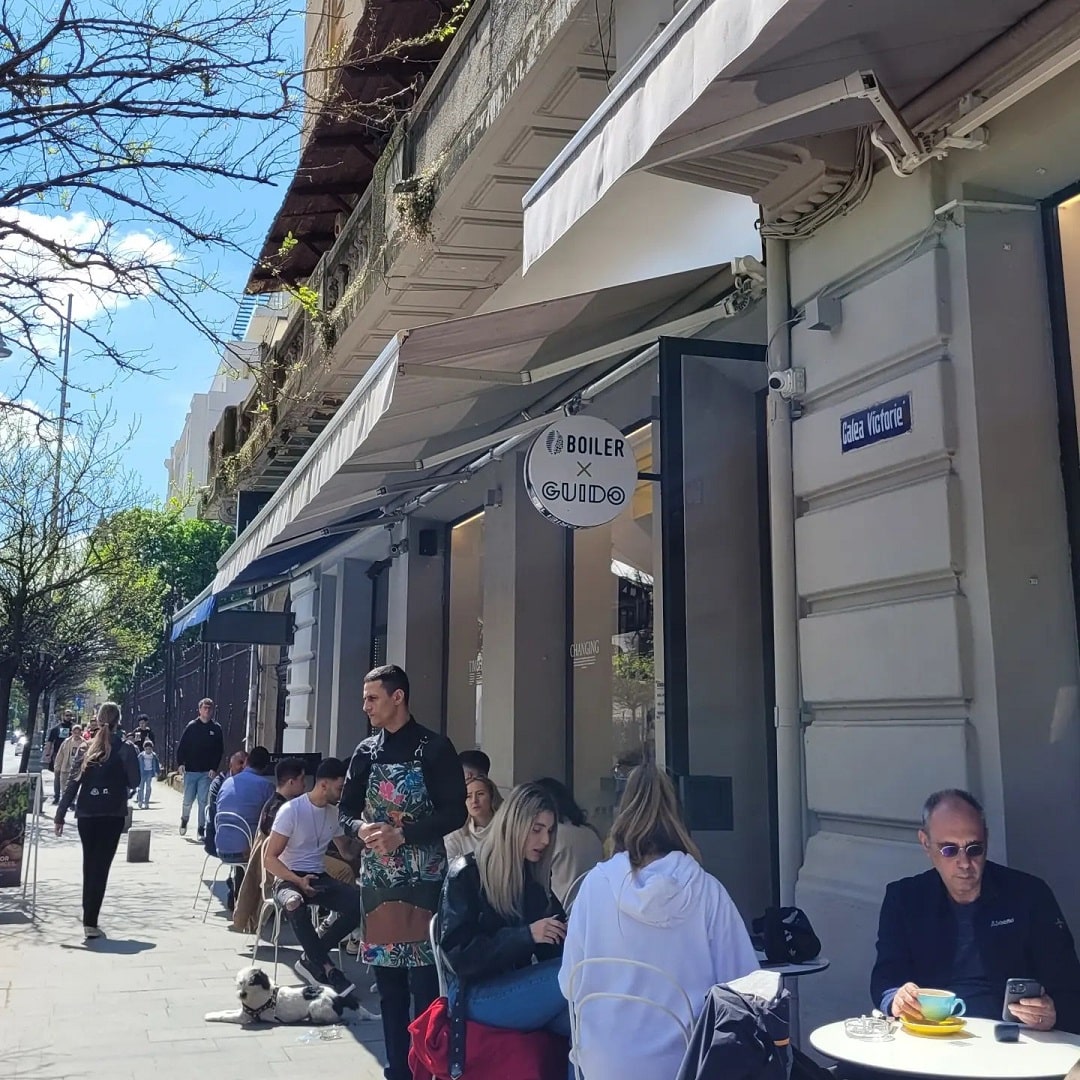 Oameni care iau masa pe terasa La bOilerXGuido la mesele mici asezate direct pe trotuar