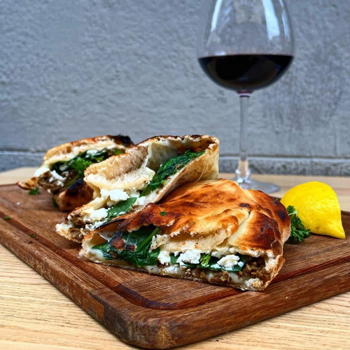 Plăcintă cu brânză și spanac taiata in felii triunghiulare, pe un tocator de lemn, langa o felie de lamaie si un pahar de vin roșiu. La Pâine și VIn. Cele mai bune tarte și plăcinte din București