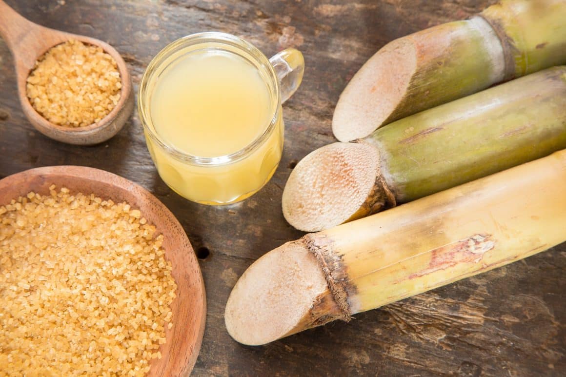 close up cu trestie de zahăr si un castron cu zahăr din trestie, alaturi de o cană cu bauturi indulcită.