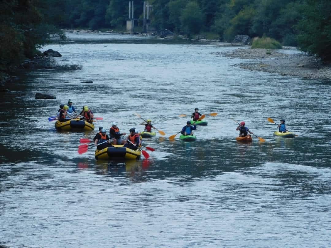 mai multe barci gomflabile si caiace cu oameni in ele plutesc pe un rau agitat, care trece printr-o padure