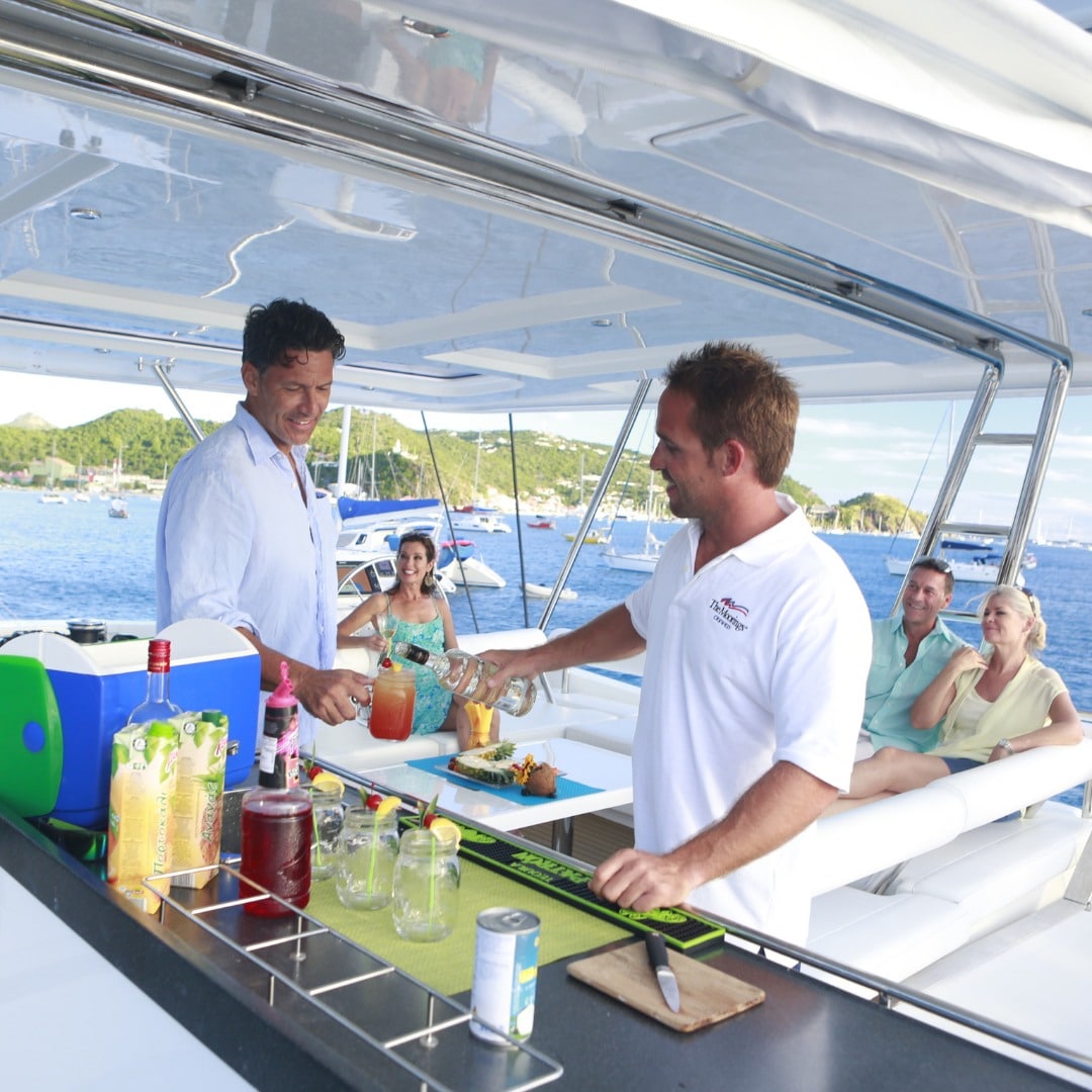 doi barbati tineri imbrcati in alb prepara cateva bauturi pe puntea unui iaht, sub prelata alba