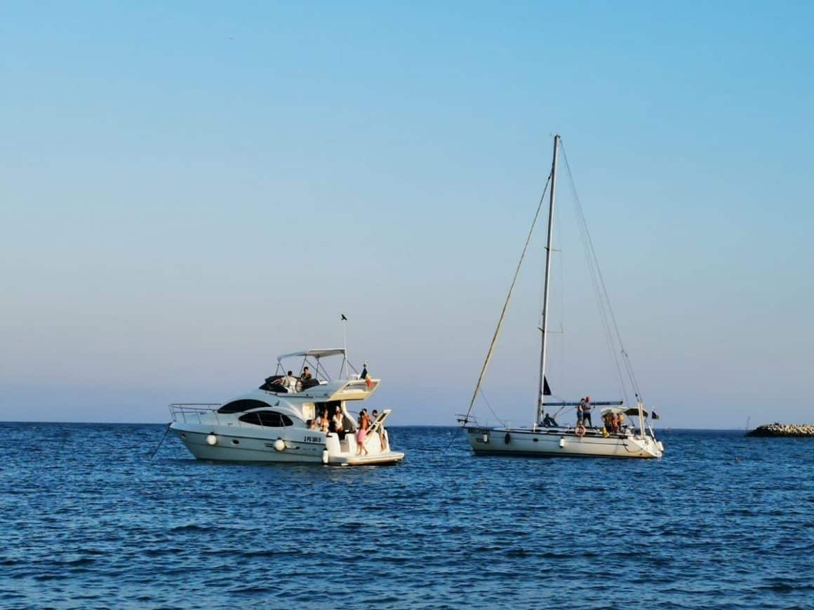 un iaht și un velier cu panzele stranse și oameni pe punte putesc pe marea calma