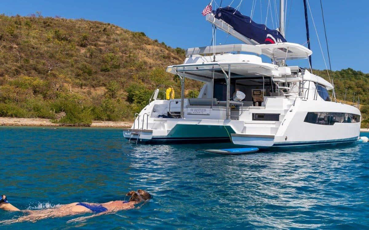 o femeie in costum de baie face snorkeling in spatele unui catamarancu panzele stranse intr-un golf din marea mediterana