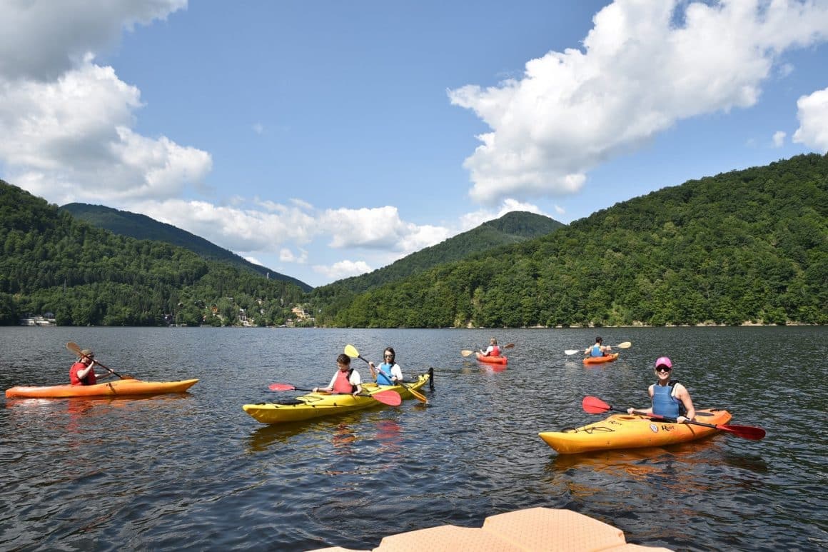 Mai multe persoane in caiace, pe lacul Tarnița din Cluj, iar in fundal muntii impaduriti, vara. Destinații de vară pentru familiile cu copii