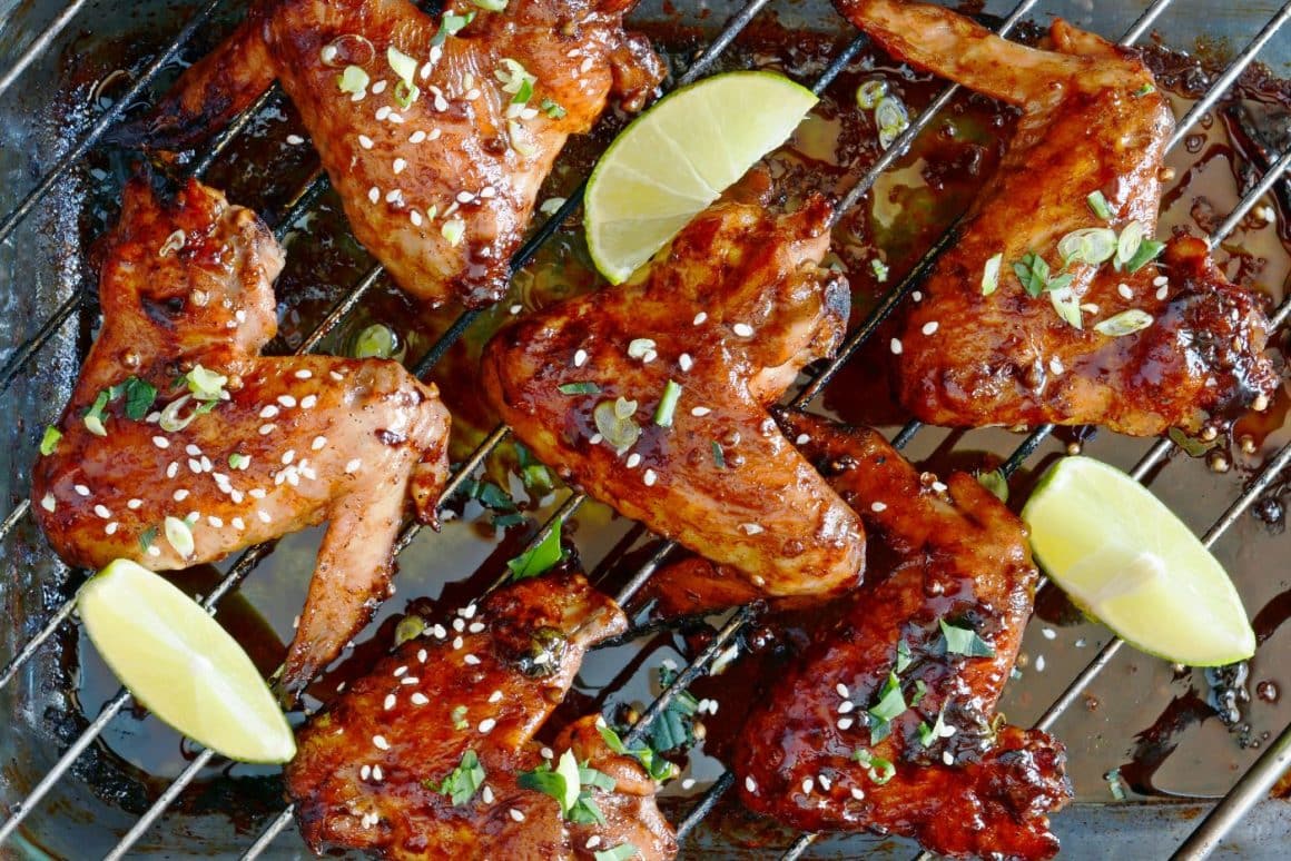 mai multe aripioare picante sunt asezate pe un grătar și sunt ornate cu verdeată tocată, susan și felii de lime