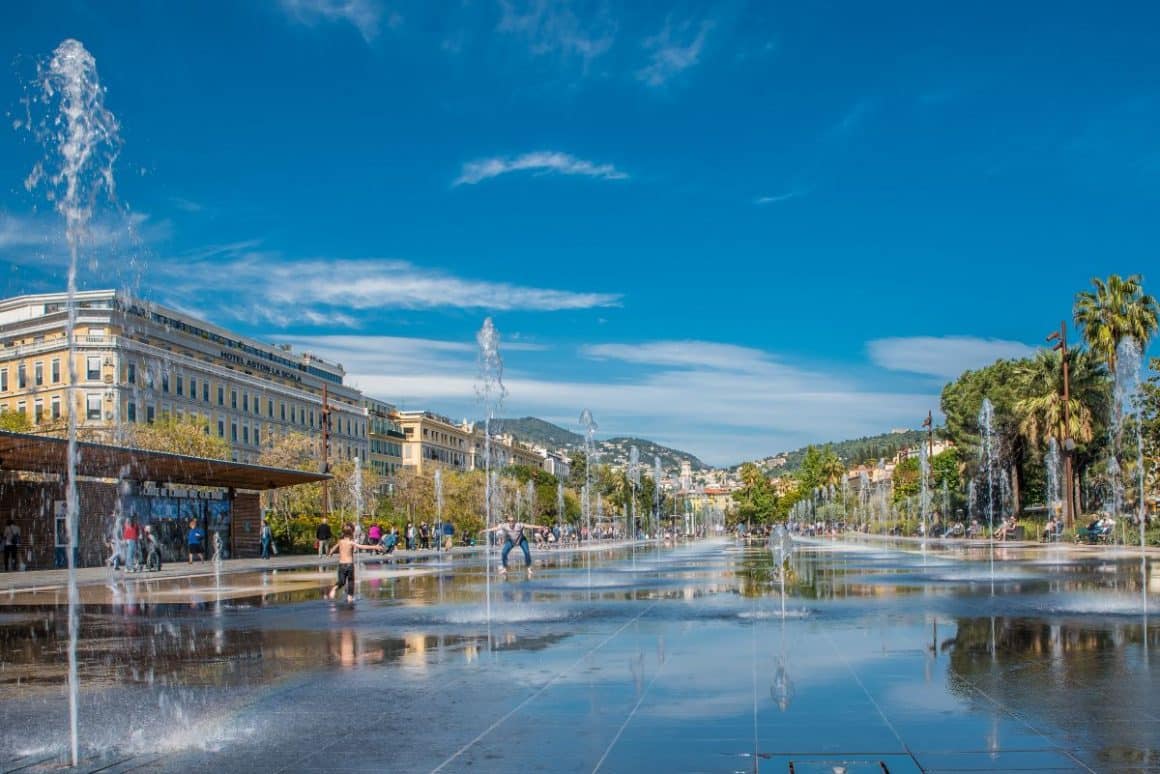 mai multe fantani arteziene sunt fotografiate pe unul dintre bulevardele orasului nisa. mai multi copii se joaca printre jeturile de apa