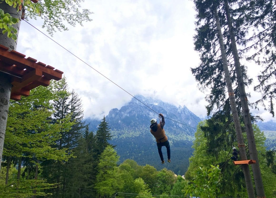 un copil coboara pe tiroliana printre mai multi copaci, iar in fundal se vede un munte stancos. 