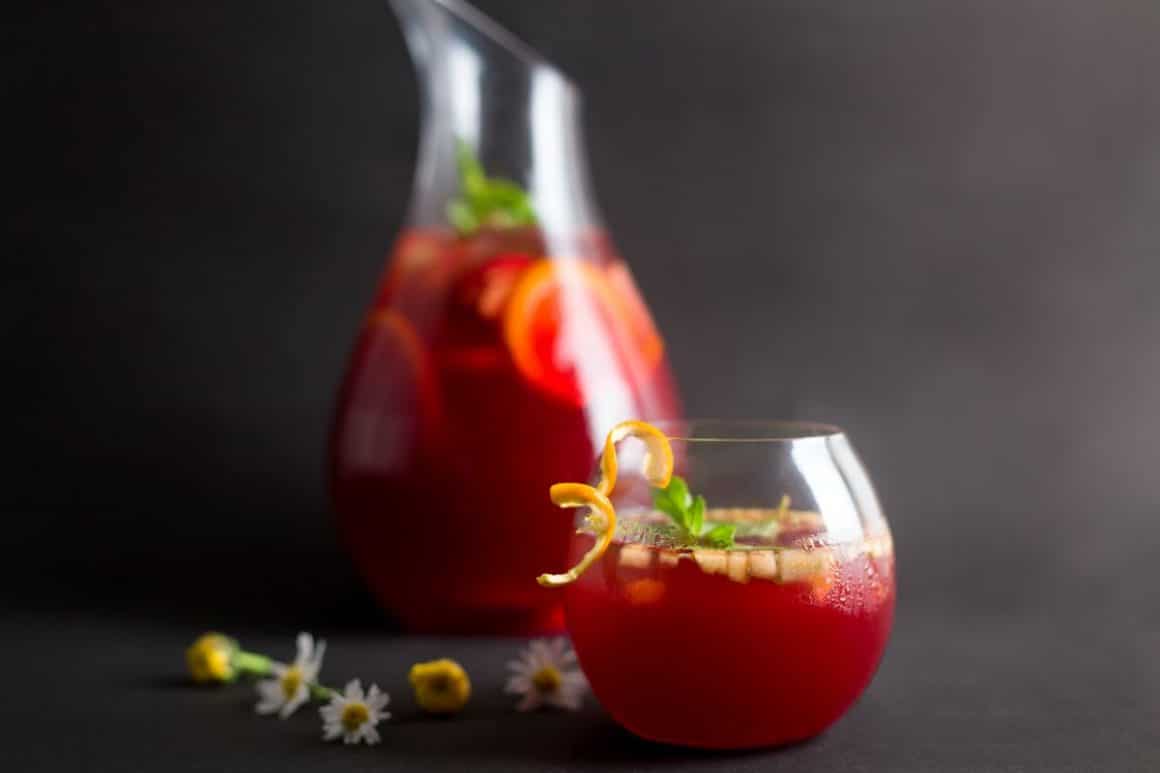 un pahar de sangria decorat cu ierburi aromatice, flori și coajă de lămâie stă în fața unei carafe cu sangria, pe un fundal gri