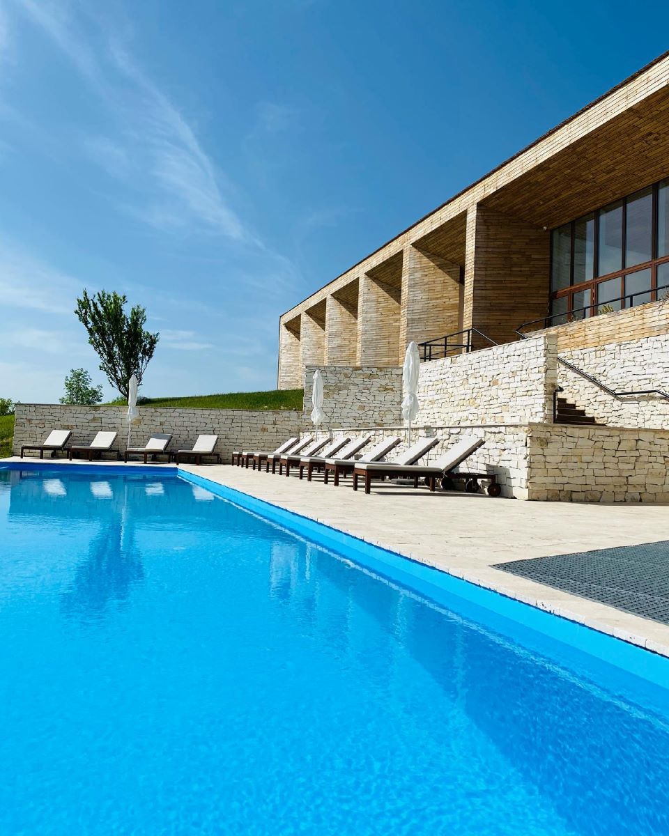 o piscina mare cu sezolnguri cu perne albe, în fata unei clădiri mdoerniste din caramida și lemn