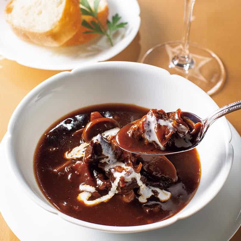 o lingură plină de mancare este ridicata deasupra unui bol cu o tocană, iar langa este un pahar și o fafurie cu pâine