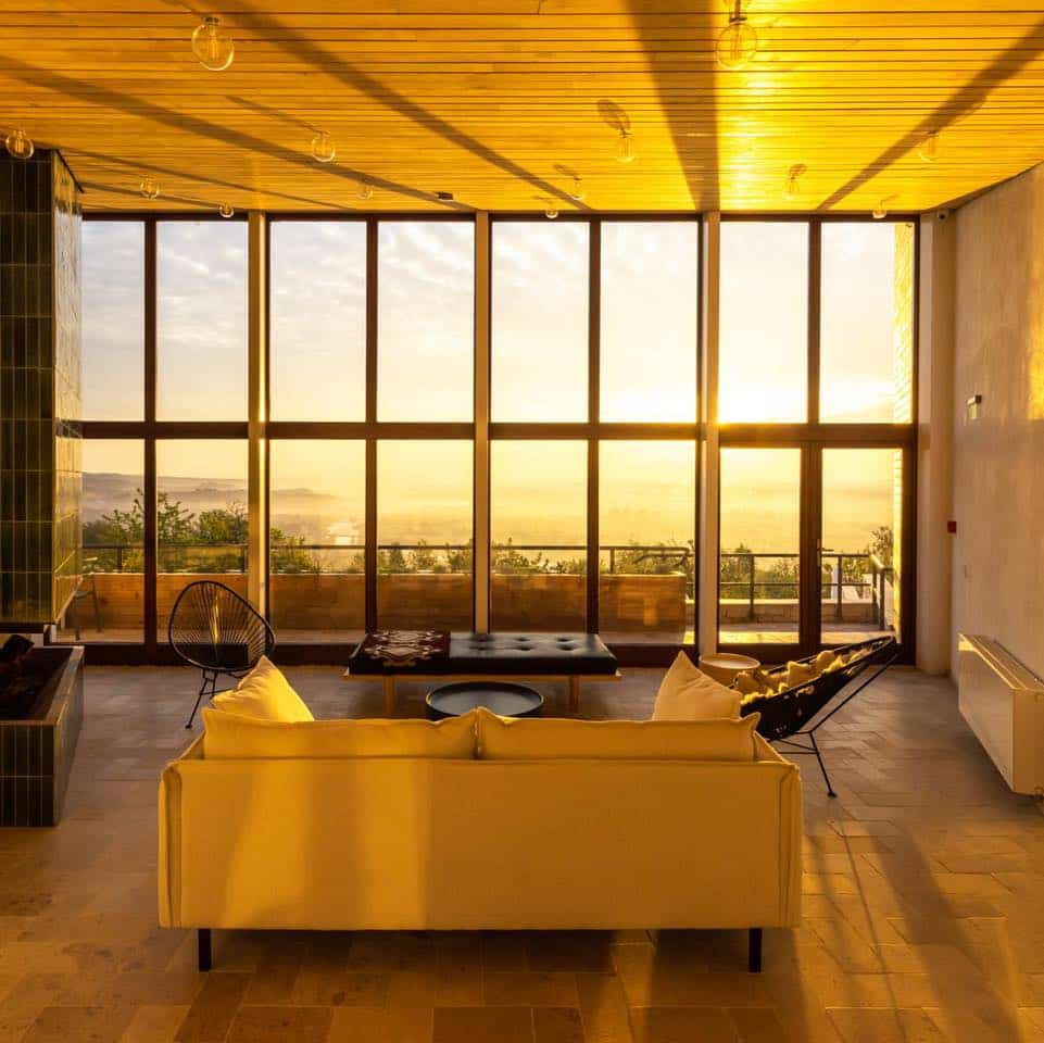 interiorul vilei cheia cu o canapea, doua scaune și o masa este foytografiat ăn lumina de apus prin geamurile mari, deaspura unei văi impădurite