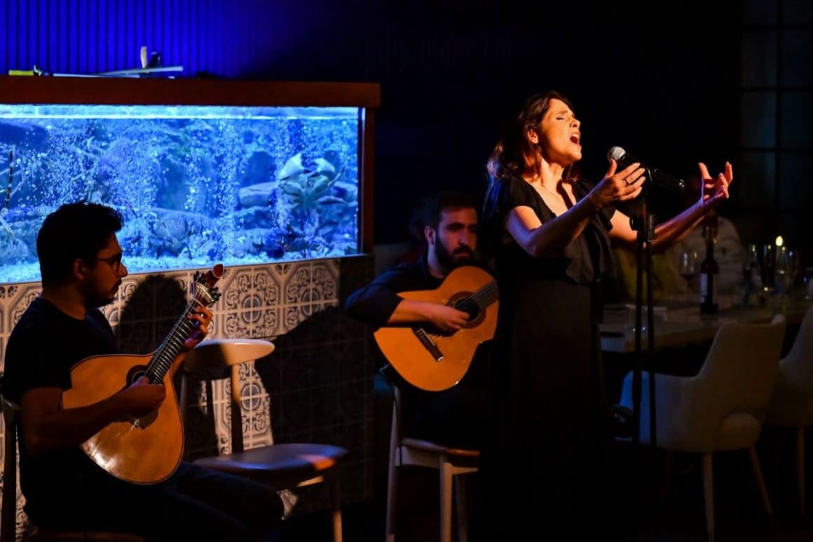 o femeie imbracata intr-o roche rosie canta la microfon pe o scena, iar in spatele ei sunt cativa instumentisti