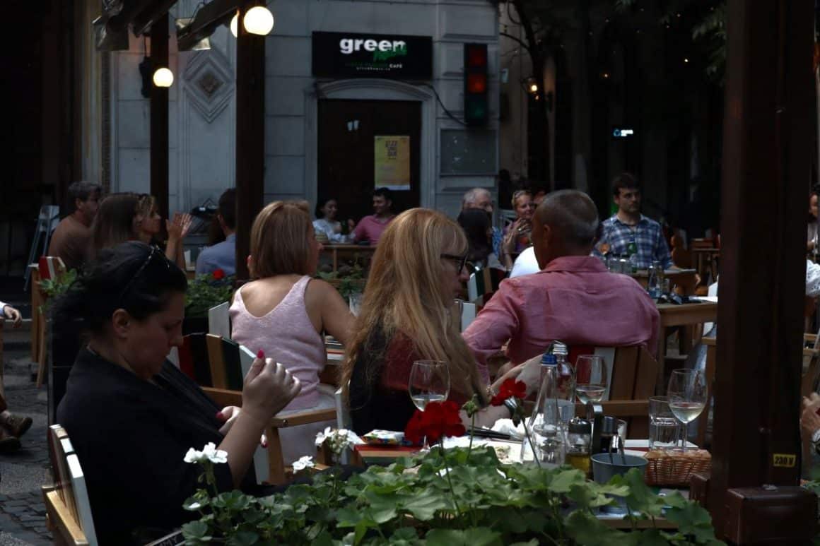 mai multi tineri stau pe scaune la una dintre mesele restaurantului Green hours, unul din locurile cu muzică live din bucuresti, in timp ce un grup de muzicieni canta pe scena