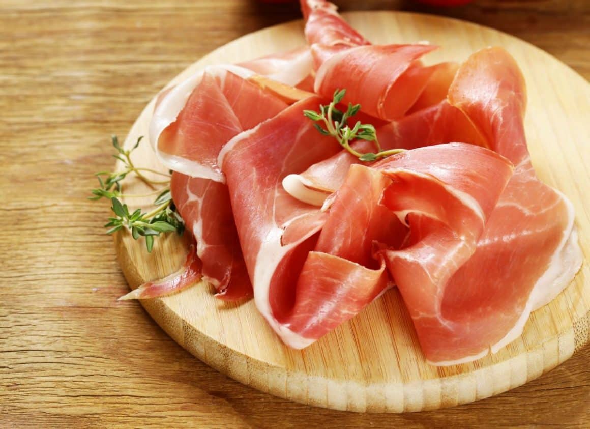mai multe felii de jamon iberico și cateva fire de verdeata sunt asezate pe un tocător de bucatarie circular din lemn, asezat pe o masă de lemn. Diferența între jamon și prosciutto