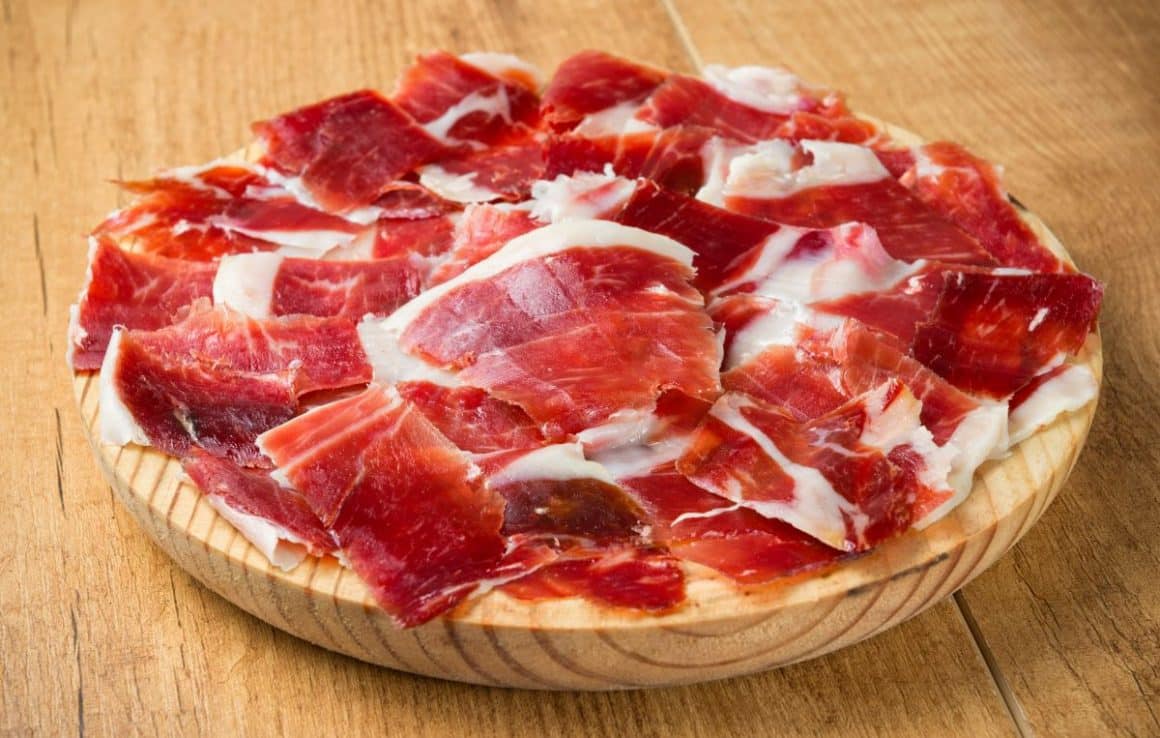 mai multe felii de jamon iberico sunt asezate pe un tocător de bucatarie circular din lemn, asezat pe o masă de lemn