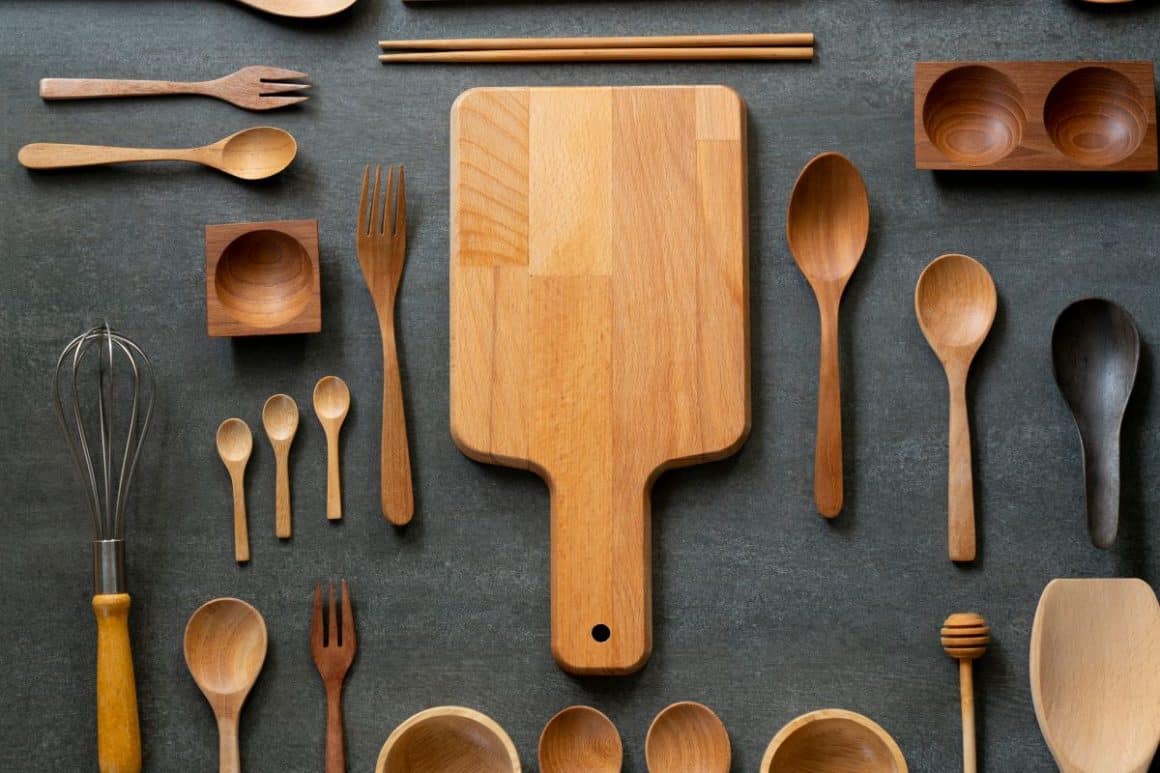 ustensile esențiale într-o bucătărie facute din lemn. mai multe linguri, un tocător și cateva boluri toate de lemn sunt asezate pe un fundal gri